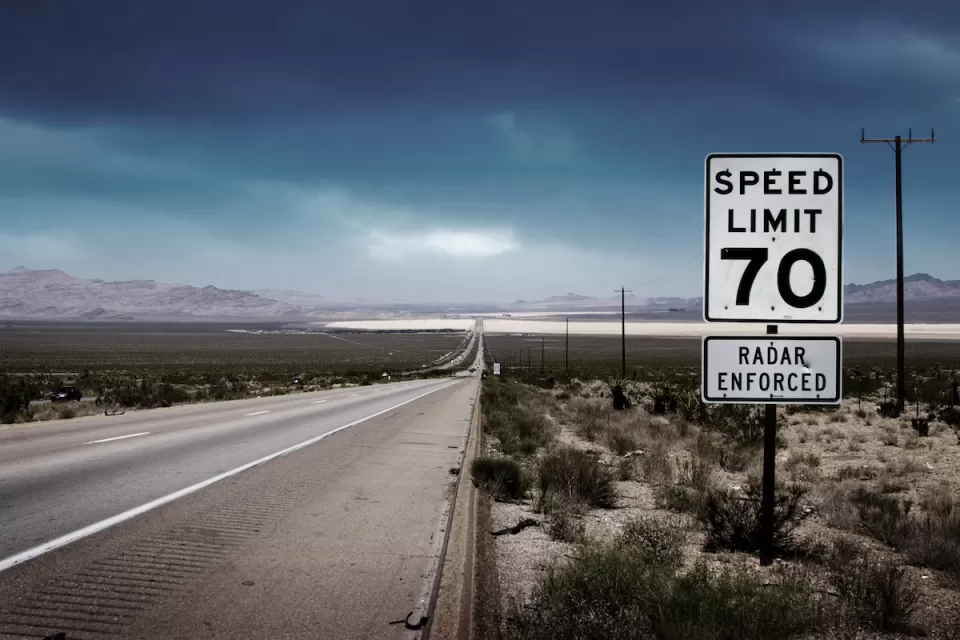 70 mph speed limit sign with bullet holes on southwestern highway with desert mountains in background.